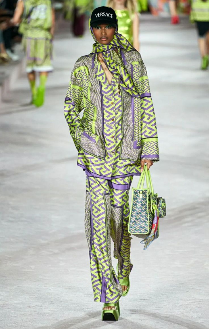 a model walks the runway during a fashion show, showcasing a vibrant lime green and purple patterned pantsuit.  the outfit is pajamastyle, loose and oversized, and features a matching scarf and versace baseball cap.  the model also carries a patterned handbag and wears platform sandals.  the background is blurred, showing other models and audience members. the overall aesthetic is one of high fashion and luxury.