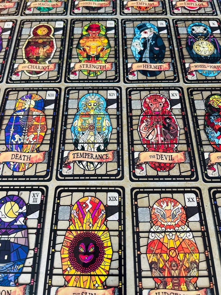a closeup image shows a collection of tarot cards laid out in a grid. the cards have a stained glassinspired design, with each card depicting a different major arcana archetype in this style. the cards are labeled with their names and roman numerals. some visible cards include "the magician," "high priestess," "empress," "emperor," "the chariot," "strength," "the hermit," "wheel of fortune," "death," "temperance," "the devil," "the moon," "the sun," and "judgement." the layout and design suggest a tarot deck made to look like stained glass windows.