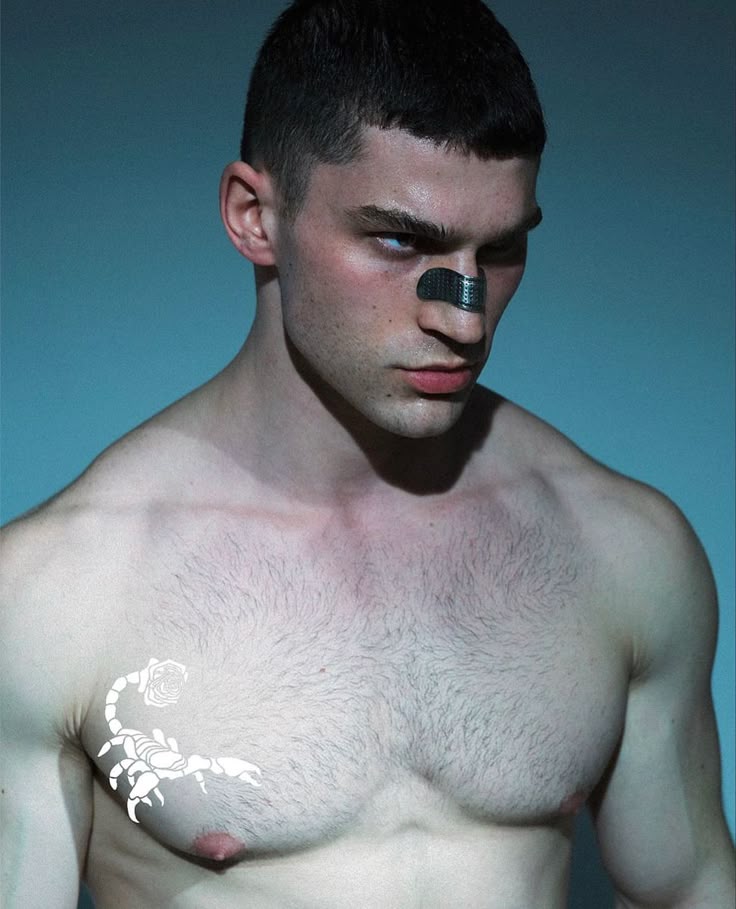 a barechested man with a scorpion tattoo on his chest and a black strip on his nose stands against a blue background. he has short, dark hair and a muscular physique.