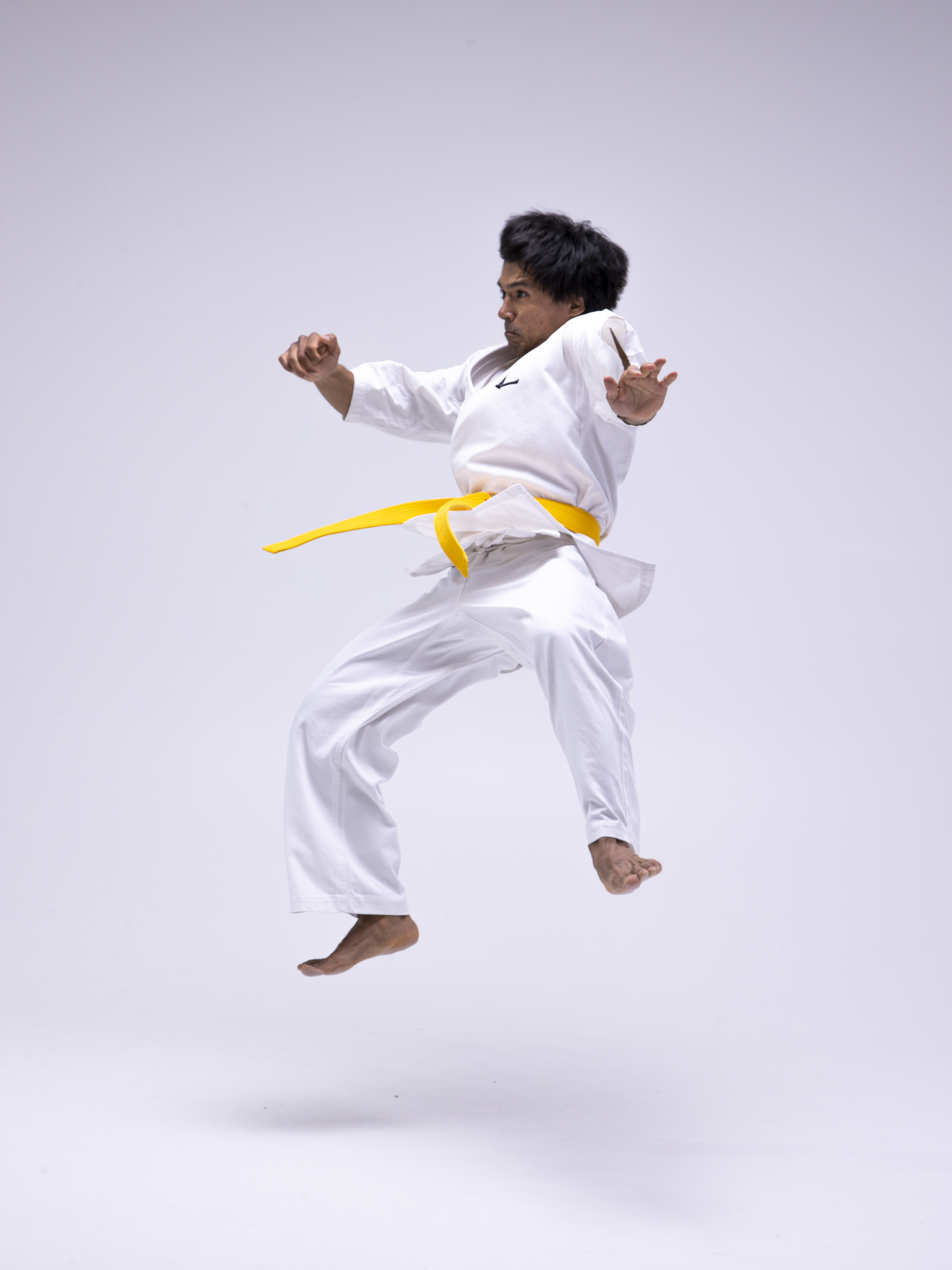 a man in a white karate uniform with a yellow belt is captured midair performing a karate move against a white background in a studio setting. he is barefoot.