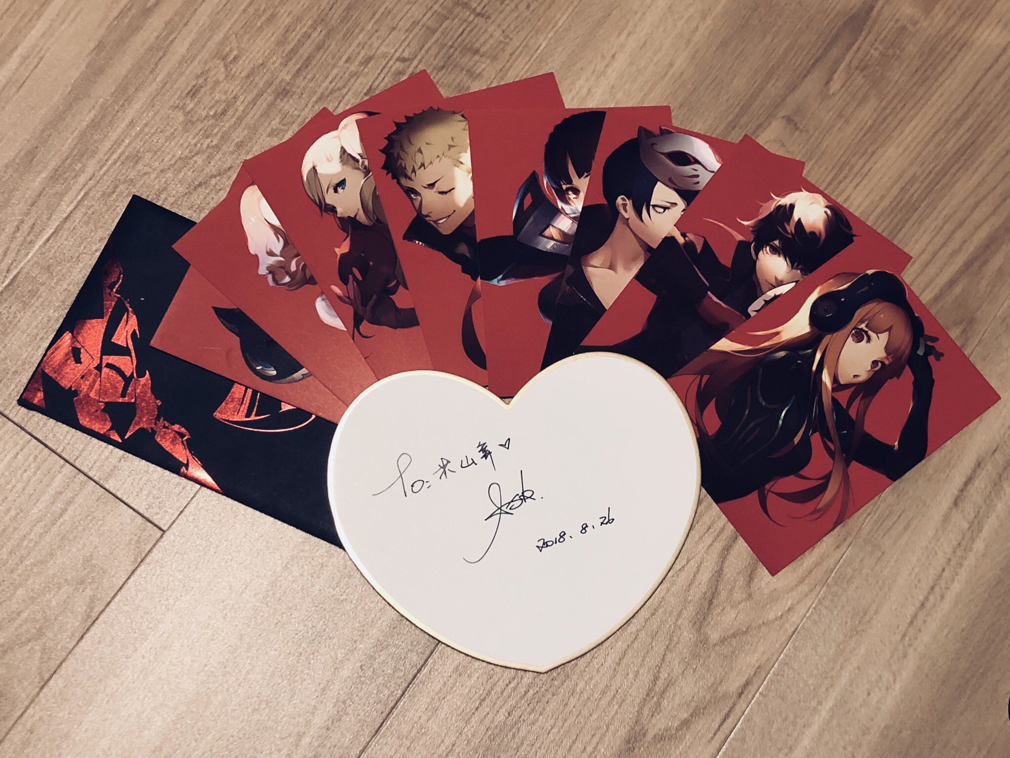 this image showcases a collection of persona 5 postcards spread out on a wooden floor.  the postcards feature individual illustrations of the main phantom thieves characters against a red background. in the center lies a white heartshaped object, likely a shikishi (japanese autograph board), adorned with a signature and date. the scene suggests a fan's prized possessions related to the popular video game.