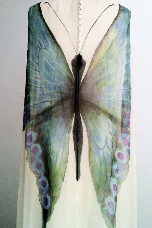 closeup shot of a white chiffon dress adorned with a large butterfly wing design. the butterfly is predominantly blue and green with violet spots near the bottom. the dress has a line of white buttons down the back.