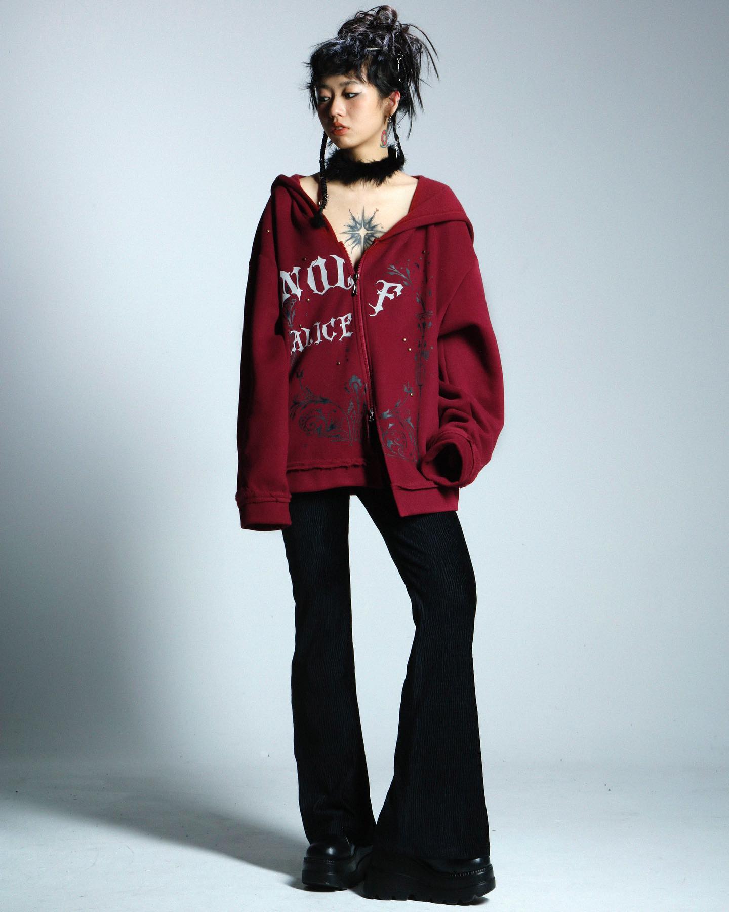 a full shot of a young woman posing in a studio against a light gray background. she is wearing an oversized burgundy hoodie with white graphic lettering, black flared pants, and chunky black platform shoes. her dark hair is styled up, and she is wearing a black choker necklace.  the overall style is edgy and alternative.