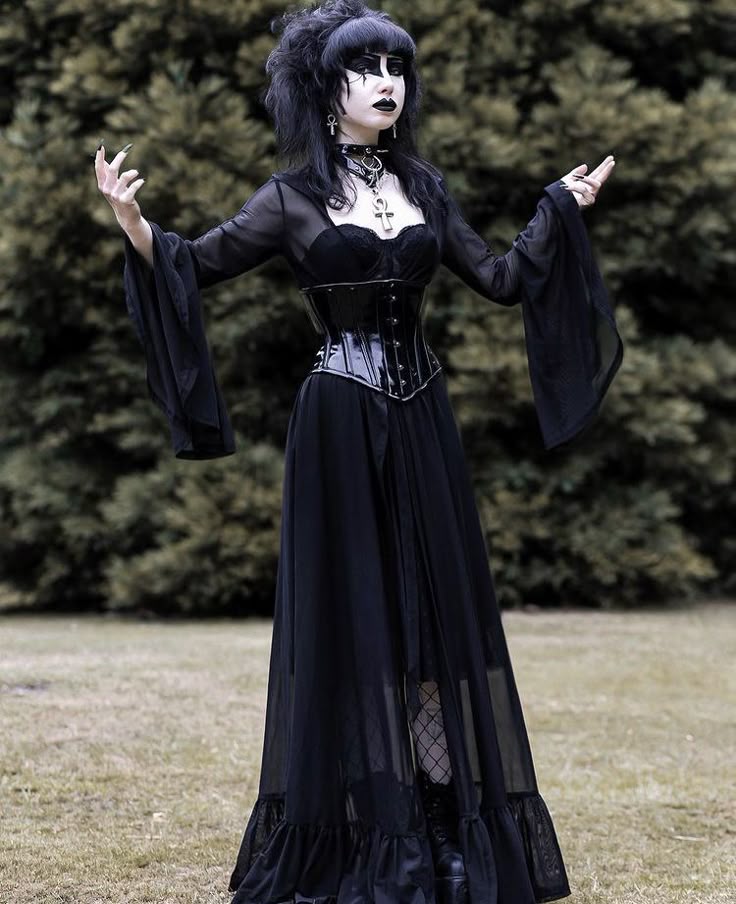 a woman in goth fashion stands outdoors. she wears a long black dress with sheer fabric and long sleeves, a corset, and boots. she also has dark makeup and jewelry. the background includes grass and trees.
