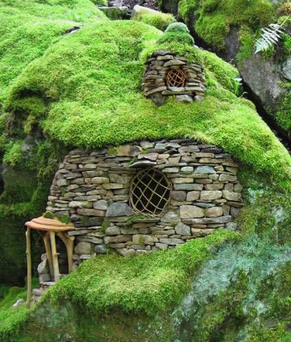 a whimsical fairy house nestled among lush green mosscovered rocks and foliage. the house is constructed from stone, featuring a small round window and a chimneylike structure on top. a miniature wooden porch is attached to the side of the house, adding to its charm and appeal. the surrounding landscape is dominated by moss and rocks, creating a natural and enchanting setting.