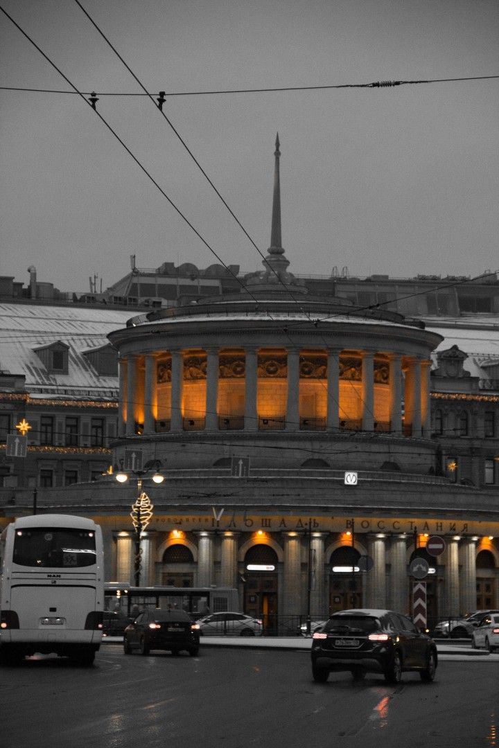 a captivating black and white photograph of the moskovsky train station in saint petersburg, russia. the image features selective color highlighting the warm, amber glow of the building's interior lights, creating a striking contrast against the cold, gray tones of the winter cityscape.  the architectural details of the station are clearly visible, showcasing its classical design and imposing columns.  trolleybus wires stretch across the sky, adding to the urban atmosphere.  several cars and a bus are visible on the street in front of the station, adding to the scene's realism and scale. the overall mood is serene yet dynamic, capturing the beauty of the city in a unique way.