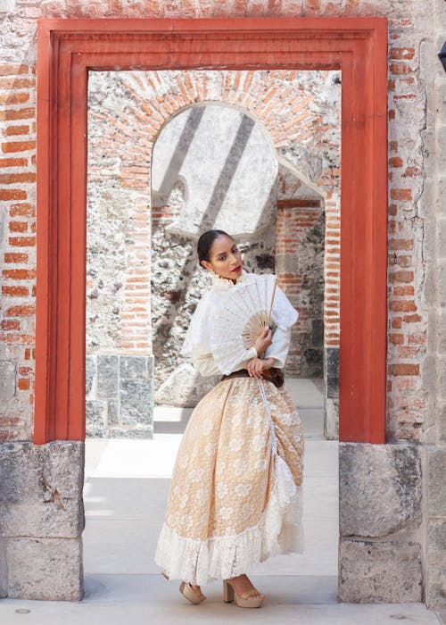 a fulllength portrait of a woman in a beautiful beige lace dress and a white lace shawl. she holds a hand fan and stands in a doorway of a colonialstyle building with brick and stone architecture, creating a captivating contrast between her elegance and the rustic background. the sunlight and shadows add depth to the image.