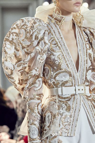loseup shot of a model showcasing a stunning white gown adorned with intricate gold embroidery and sequins. the dress features dramatic puff sleeves, a belted waist, and a deep vneckline. the fabric appears to be sheer, allowing the underlying details to shine through. the model is wearing statement gold earrings that complement the dress’s embellishments. the overall aesthetic is one of high fashion, luxury, and elegance. the background is blurred, keeping the focus on the dress and its exquisite detailing.