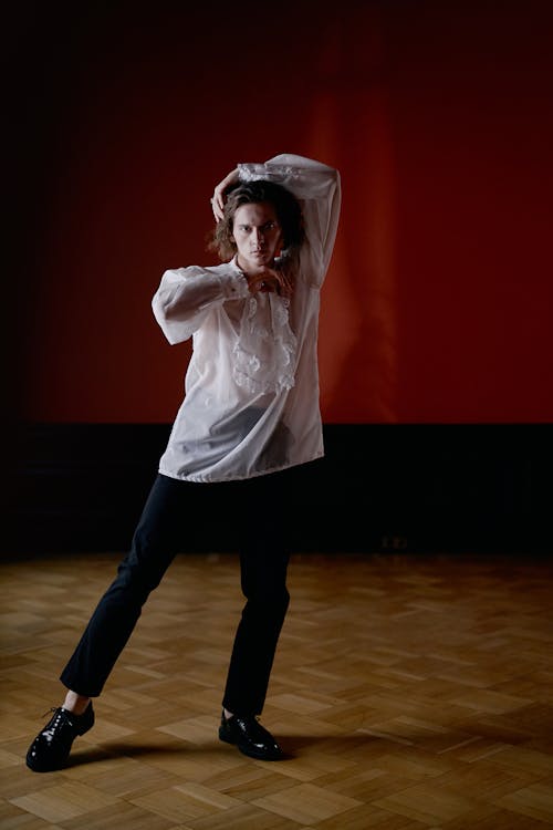 the image shows a young man, possibly a dancer, in a white, sheer shirt and black pants. he's posing dramatically against a dark red wall in a studio with a wooden floor. his posture suggests movement and expression; his hands are raised to his head, and his body is slightly angled.  the overall mood is artistic and theatrical. the lighting highlights him against the dark background. he is wearing black shoes, and his hair is long and dark. the image has a classical or contemporary dance feel.