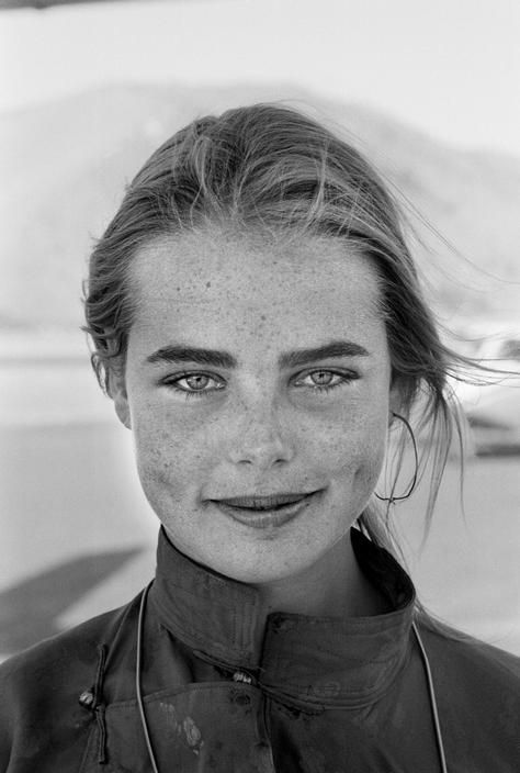 a black and white portrait of a young woman with freckles. she is facing the camera with a slight smile. her hair is windblown and she is wearing a collared shirt and a single hoop earring. the background is blurred, suggesting an outdoor setting with natural light.