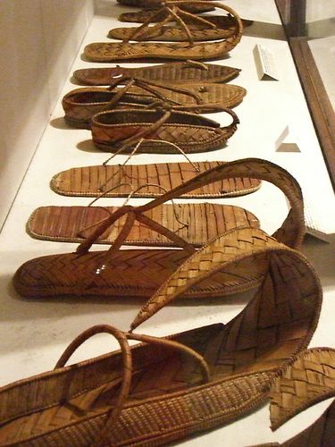the image shows a display case with several pairs of traditional woven sandals made of straw or a similar material. the sandals are displayed in a row, allowing viewers to see the details of their construction and design. they appear to be part of a museum exhibit showcasing cultural artifacts.