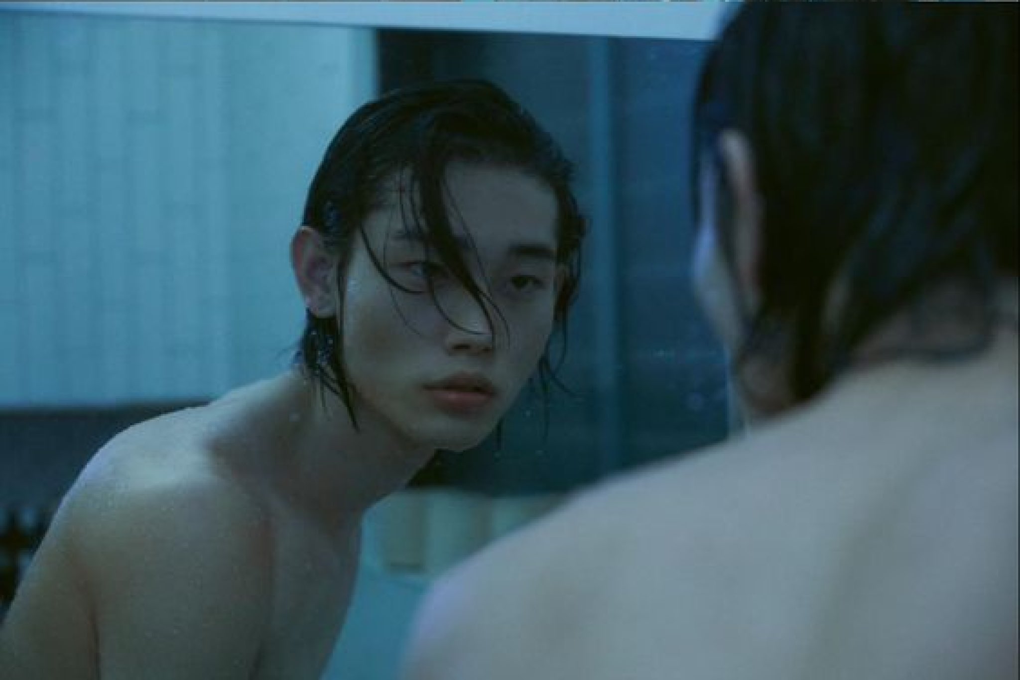 a young man with dark hair is looking at the mirror with a serious face. the reflection of the young man is in the mirror. the young man is wearing no shirt, his chest is wet from water drops. the background of the image is blue toned. the young man appears to be in the bathroom.