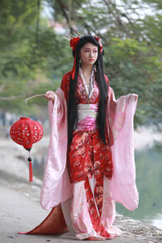 a young woman in a beautiful red and pink kimonostyle dress stands gracefully outdoors, holding a red paper lantern. her long black hair is styled traditionally, and she exudes an air of elegance and serenity. the background is blurred, focusing attention on the woman and her stunning attire. the overall image is evocative of asian culture and traditional clothing.