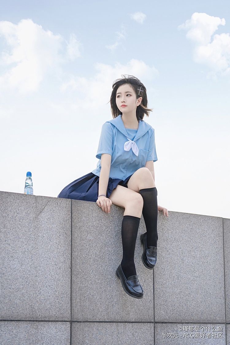 a young woman wearing a light blue school uniform with a navy blue skirt and black socks and loafers is sitting on a gray wall. she has a cross hair clip in her hair and is looking away. a bottle is sitting on the wall behind her. the sky is blue with white clouds in the background.