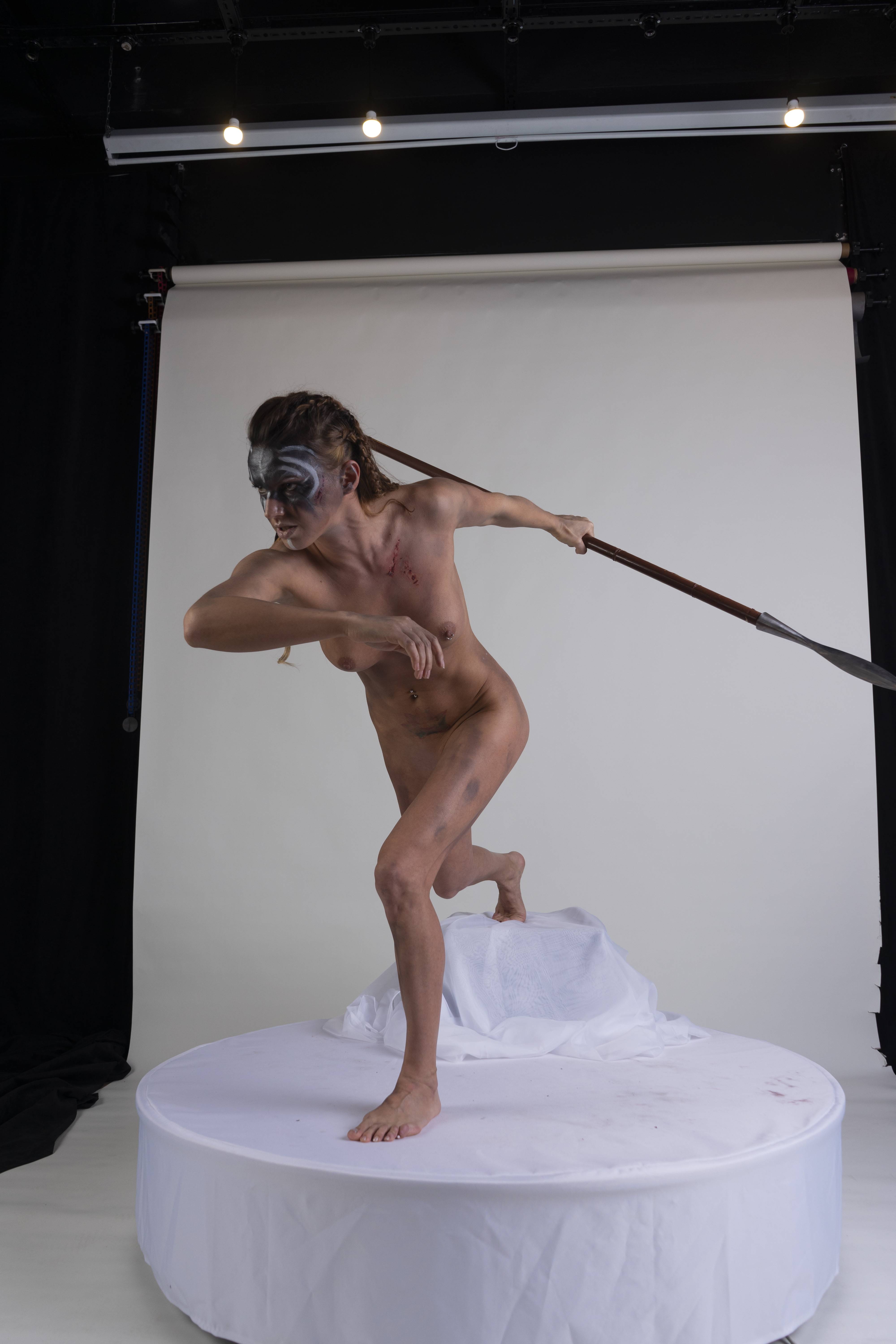 a nude woman with face paint and braids stands in a fighting stance, holding a spear. she is in a studio setting with a white background.