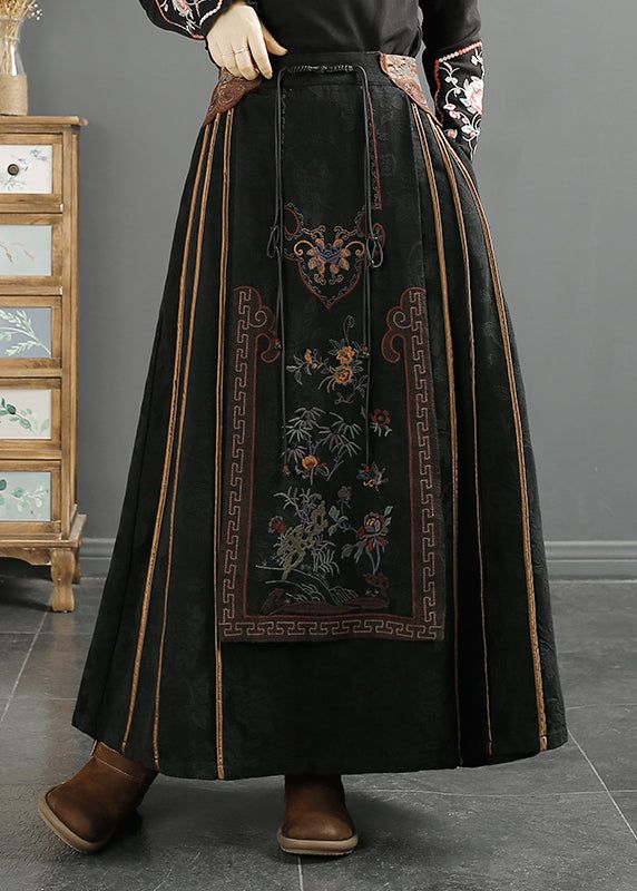 a person is wearing a black, embroidered skirt that appears to be a hanfu or other type of folk costume. the skirt has a central panel with detailed floral embroidery and a brown border with a geometric pattern. the skirt is long and pleated, with thin brown accents running down the pleats. the person is wearing brown shoes and standing on a gray floor. in the background, there is a wooden dresser or cabinet. the lighting is soft and muted, highlighting the textures of the fabric and embroidery.