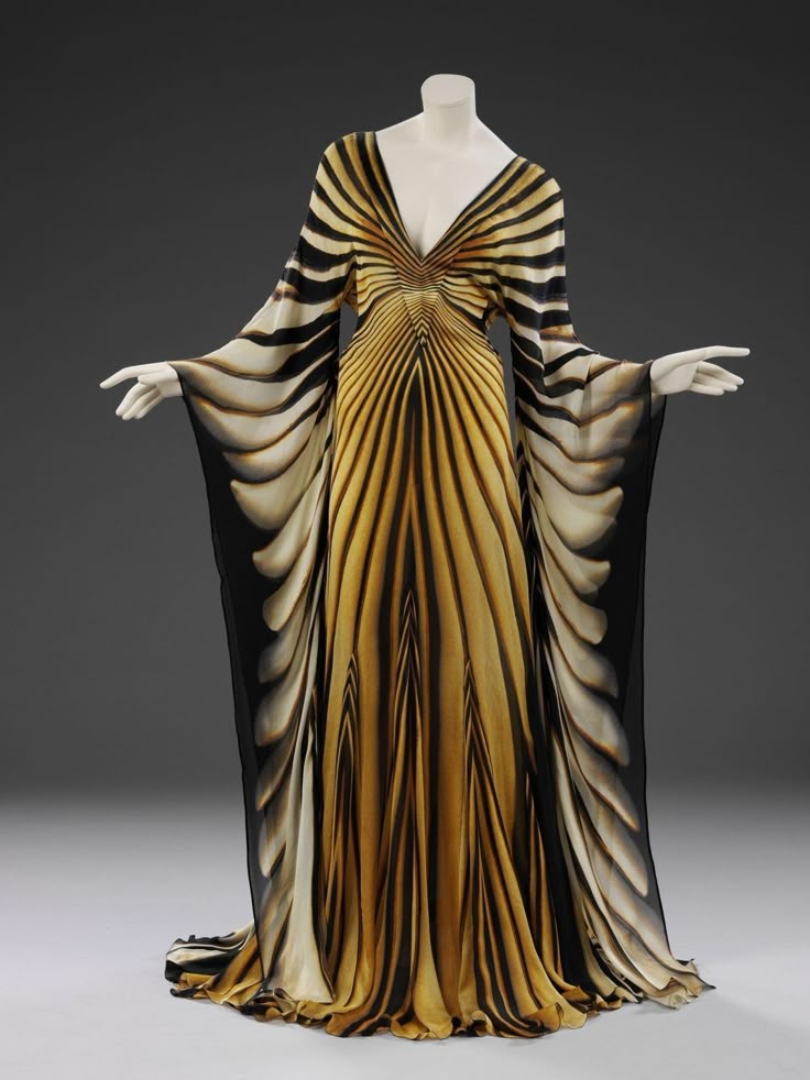 a long, flowing gown with a deep vneck is displayed on a mannequin. the dress has long, billowing sleeves and a striking pattern of black, gold, and white stripes and curves. the fabric appears to be chiffon or a similar lightweight material, creating a draped effect.
