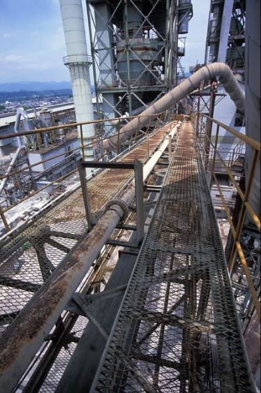 highangle, closeup view of an elevated walkway or catwalk in a large industrial factory or plant. the walkway is made of metal grating and runs along the side of a large structure, with pipes and other industrial equipment visible. the scene shows rusty and aged metal components, suggesting a wellused facility. the background includes other parts of the industrial complex and a glimpse of a landscape with mountains and a cityscape in the distance. the sky is mostly clear and blue, suggesting a daytime setting. the overall impression is one of largescale industrial production or manufacturing.