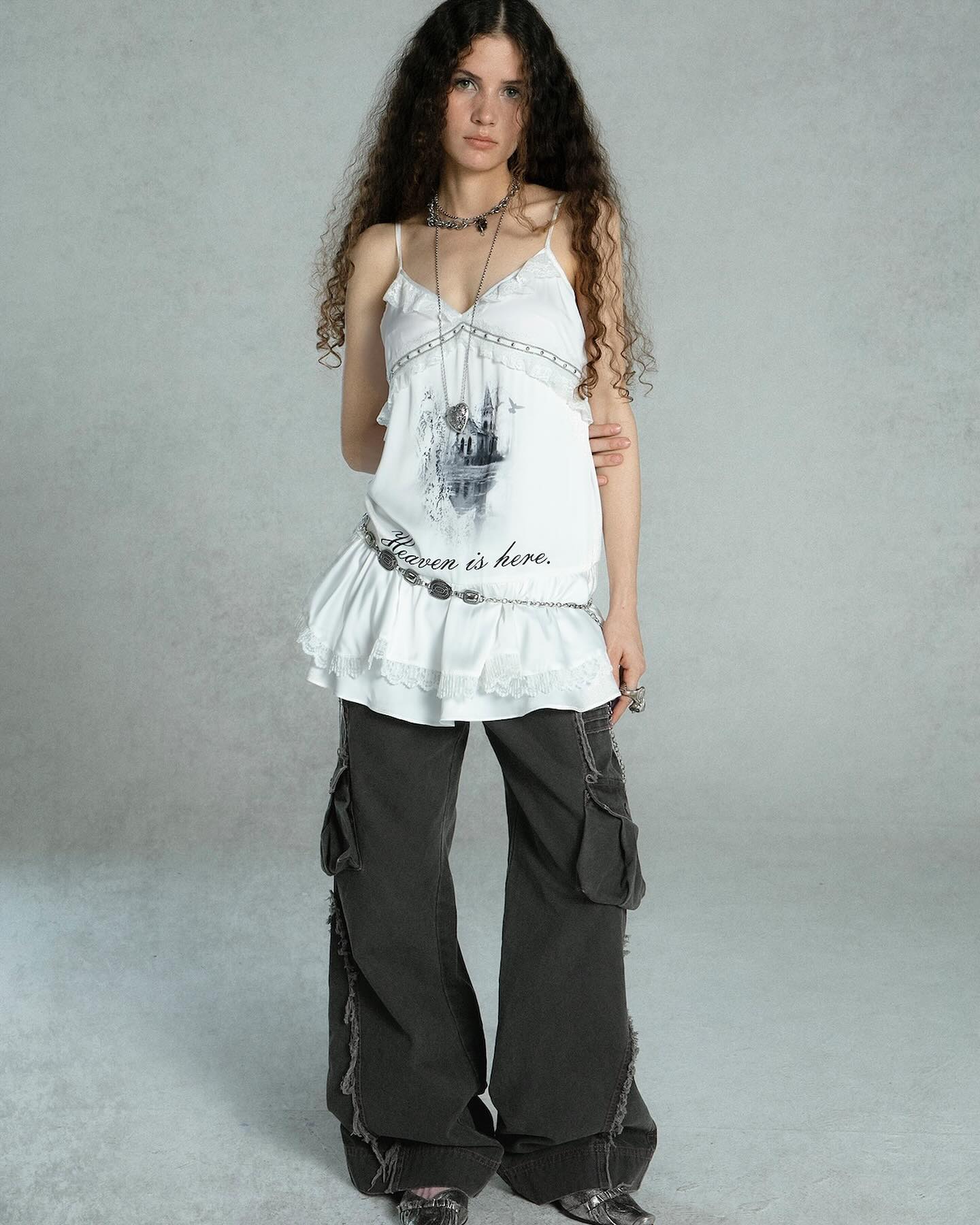 a full shot of a woman model showcasing a stylish outfit. she wears a white ruffled camisole with a graphic print and a thin lace trim. the camisole is paired with dark grey cargo pants with frayed hems. the model also wears various silver jewelry including necklaces and a belt. her long curly hair is loose and her makeup is minimal.  the background is a simple, light grey studio backdrop.