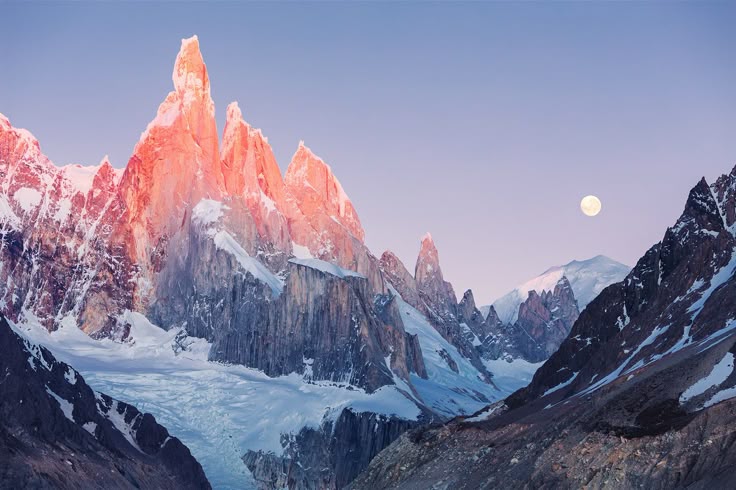 the image features a stunning mountain range at either sunrise or sunset, with the peaks of the mountains illuminated by a warm, reddish light. snow and glaciers cover the lower parts of the mountains. a full moon is visible in the sky. the sky is a light blue or pink hue, and the landscape is rugged and mountainous.
