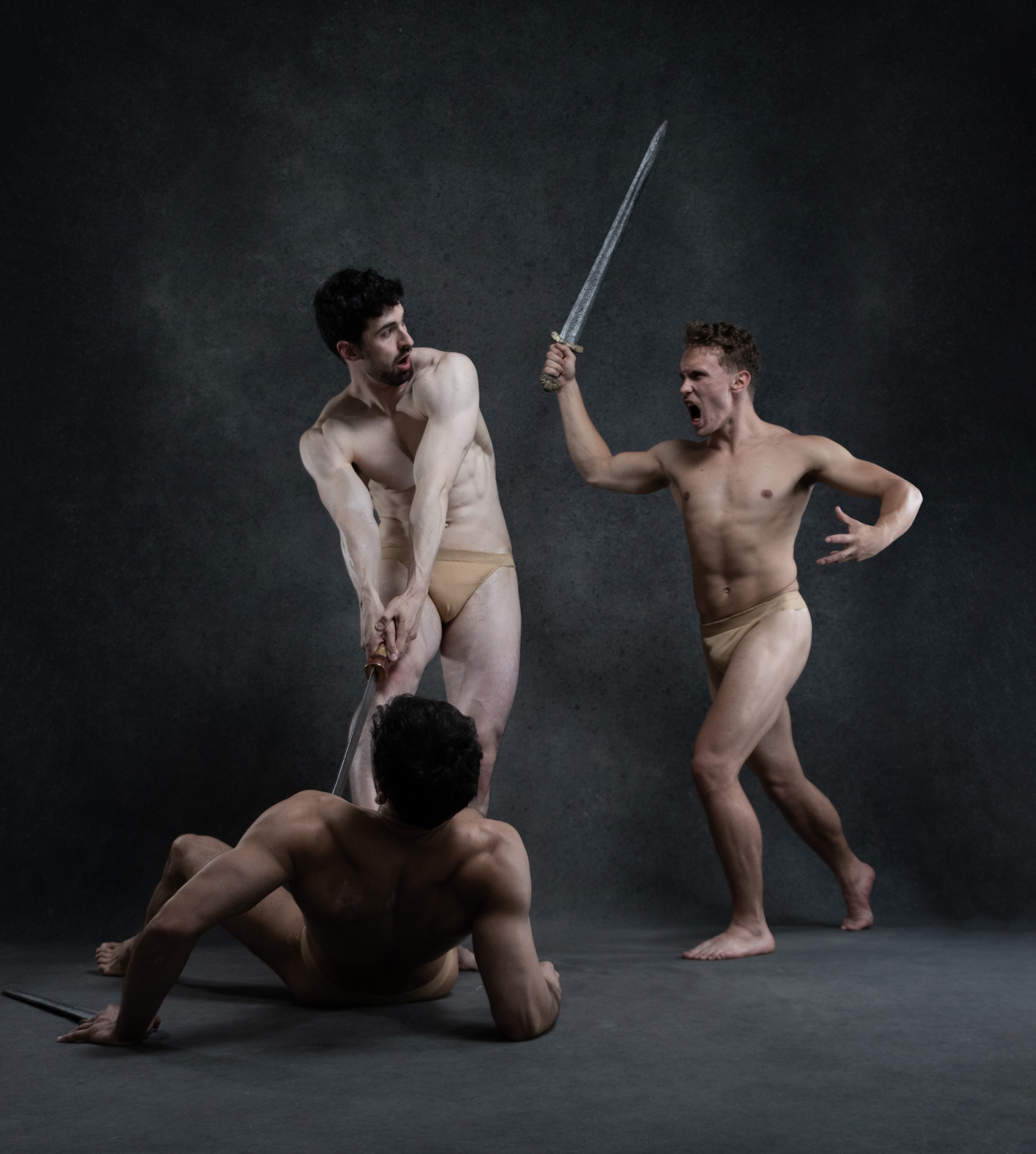 the image showcases three men engaged in a fierce sword fight set against a stark, dark grey studio backdrop. two of the men are actively engaged in combat, one wielding a sword while the other attempts to disarm him. the third man lies wounded on the ground, his sword fallen aside.  their bodies are nude, highlighting their muscular physiques and the intensity of their struggle. the studio lighting casts dramatic shadows, further amplifying the raw aggression and power dynamics of the scene.