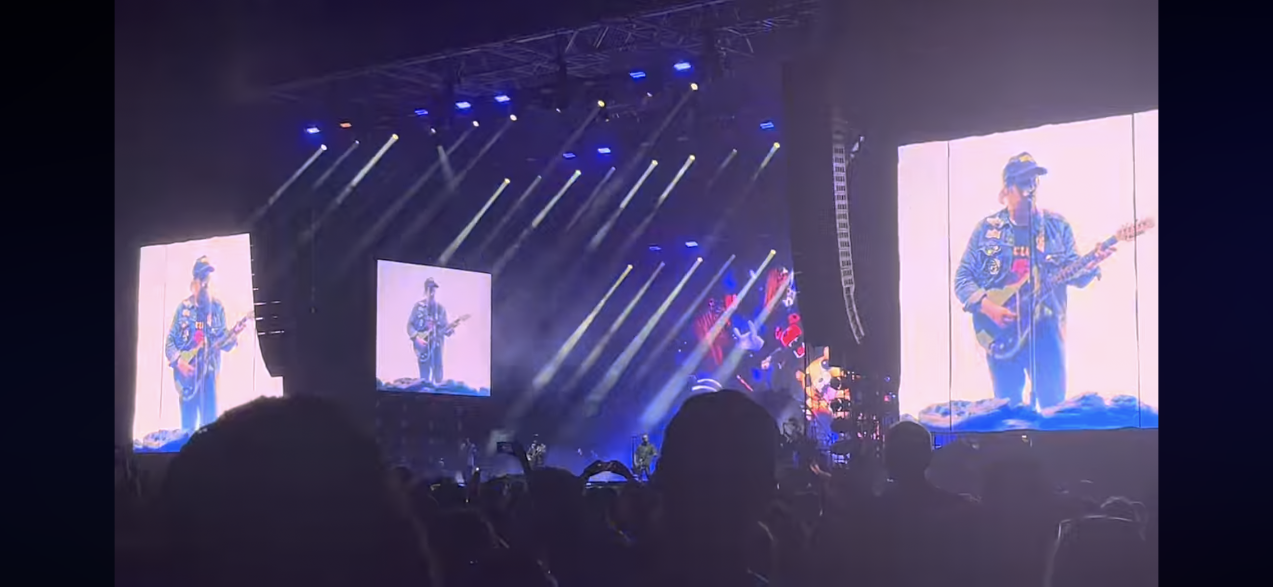 a concert stage with a large screen displaying the silhouette of a musician holding a guitar. the screen is surrounded by a crowd of people watching the performance. the stage is brightly lit with spotlights. the musician is wearing a blue denim jacket and blue jeans and a cap.