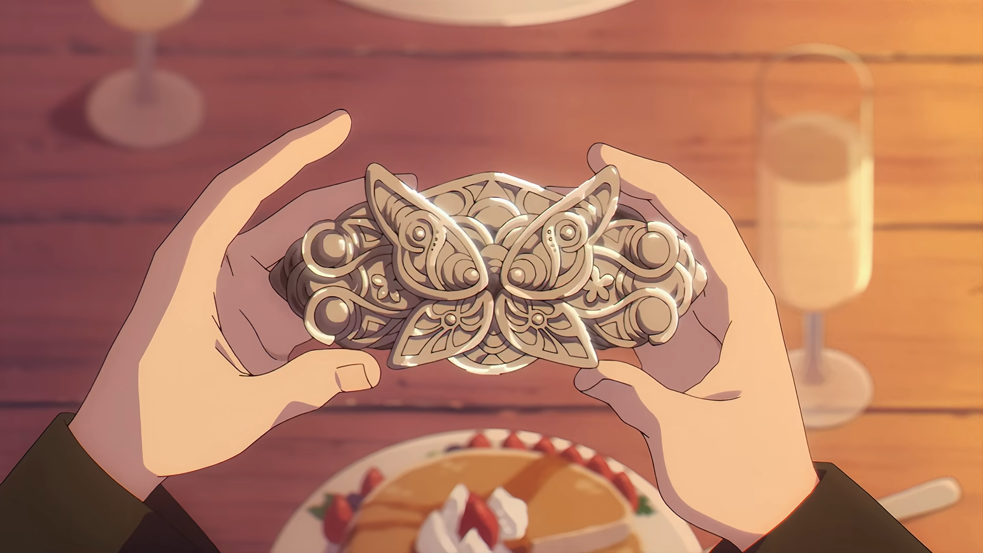 the image shows a pair of hands delicately holding a silver, ornatelydesigned butterfly ornament over a wooden table. the ornament appears to be a clasp or centerpiece with intricate details and small pearllike accents. below the hands, the scene suggests a romantic meal for two. we see a partially eaten dessert topped with strawberries and cream, likely pancakes or waffles, and two elegant champagne glasses, one empty and the other partially full. the warm, natural lighting suggests a daytime setting, creating a cozy and intimate atmosphere.