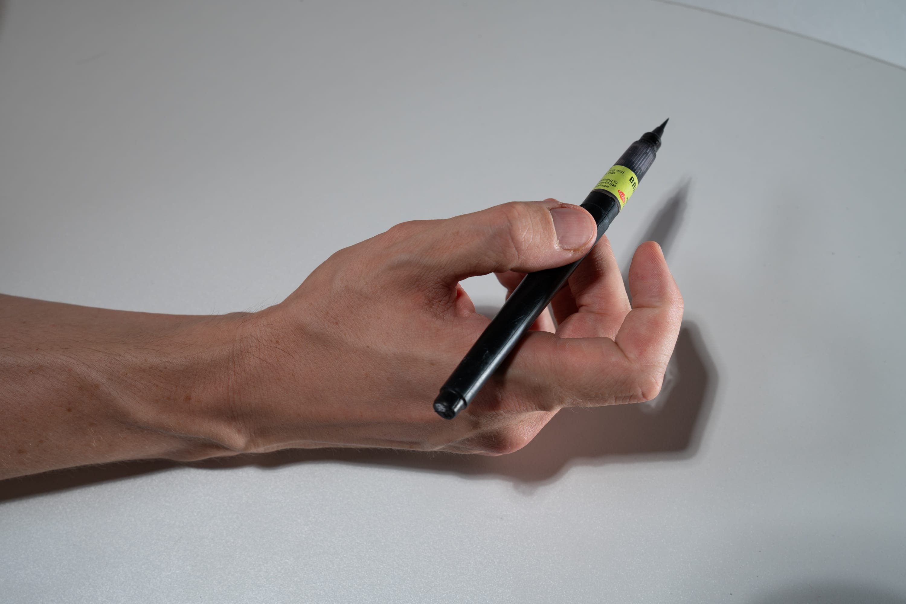 a hand holds a black brush pen against a white background. the pen is held at an angle, with the tip pointing towards the right. the hand is positioned in the lower left corner of the image, with the arm extending out of frame. the background is a plain white surface. the lighting is soft and even, creating subtle shadows around the hand and pen.