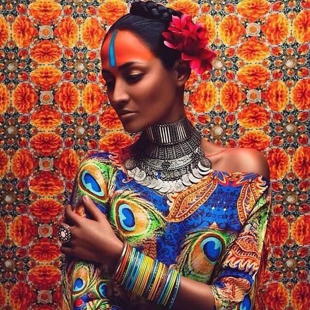 the image is a striking portrait of a darkskinned woman, possibly a model, dressed in vibrant, richly patterned clothing and adorned with elaborate jewelry. her attire seems inspired by indian or south asian traditions. she wears a blue and green paisleyprinted dress with peacock feather motifs, a large silver necklace, numerous colorful bangles on her wrists, and a decorative headpiece including a red flower.  a bindi (a decorative dot) is placed on her forehead. the background is a kaleidoscopic pattern of orange and darkred flowers creating a  mandalalike effect. the overall style is artistic and evocative of  fashion photography, possibly for a bollywoodinspired campaign or a representation of south asian culture. the woman's pose is graceful and contemplative, her gaze directed downward.