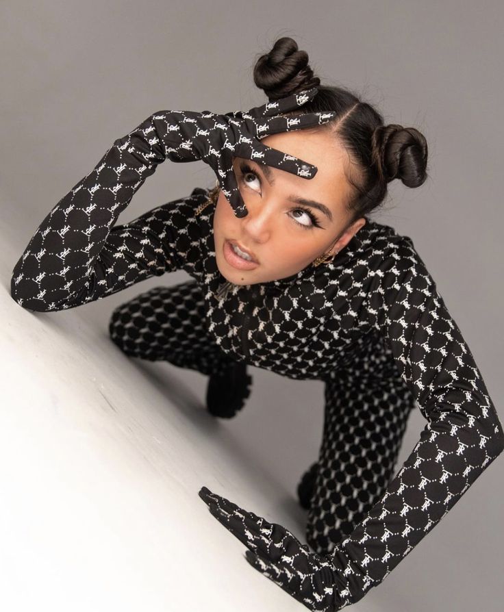 the image shows a young woman posing in a stylish black and white bodysuit. the outfit features a repeating monochrome geometric pattern. she is wearing long gloves that match the bodysuit. her hair is styled in two high buns or topknots. she's posing in a dynamic, almost crouching position,  looking upwards at the camera. the background is a plain, light gray, which keeps the focus on the model and her outfit. the overall aesthetic is modern and fashionable.
