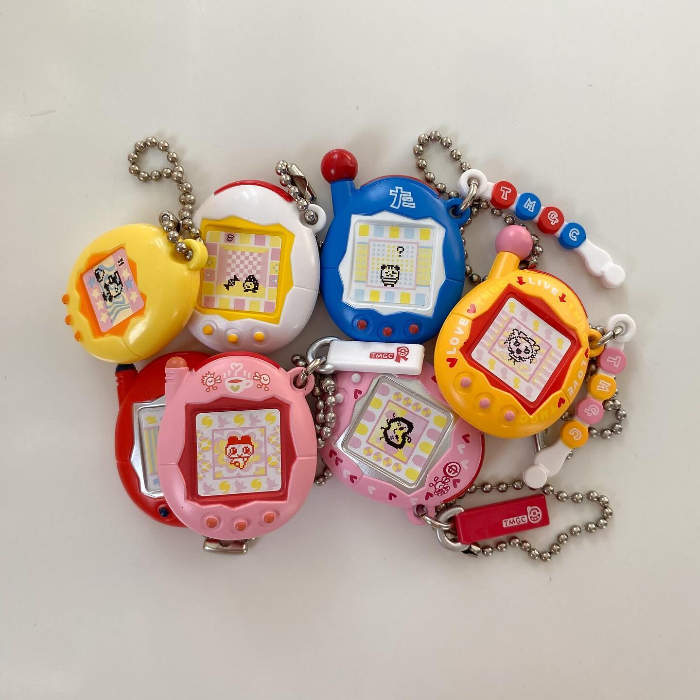 the image shows a collection of seven tamagotchi virtual pets arranged closely together on a white surface. each tamagotchi is a different color and design, featuring various characters and screen displays. the devices are attached to keychains, suggesting they are used as accessories. the style of the tamagotchis indicates they are likely vintage or retro items, reminiscent of the 1990s.