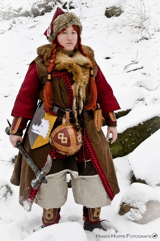 a woman in a detailed cosplay costume stands in the snow. the costume appears to be inspired by medieval or viking attire, featuring a long brown tunic with red accents, fur trim, and a hat. she is holding a battleaxe and carries several accessories like a bag and a helmet. the setting is snowy and outdoor, enhancing the authenticity of the costume.  the image is sharp and welllit.