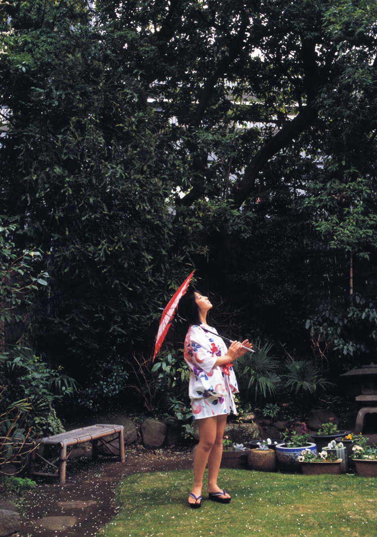 a young woman in a short white yukata patterned with pink flowers stands on a lawn and looks up at the sky. she is holding a red and white umbrella in her right hand and a small notebook in her left hand. behind her is a lush green garden with a variety of plants and a wooden bench. the sky is overcast, and the lighting is soft and diffused.