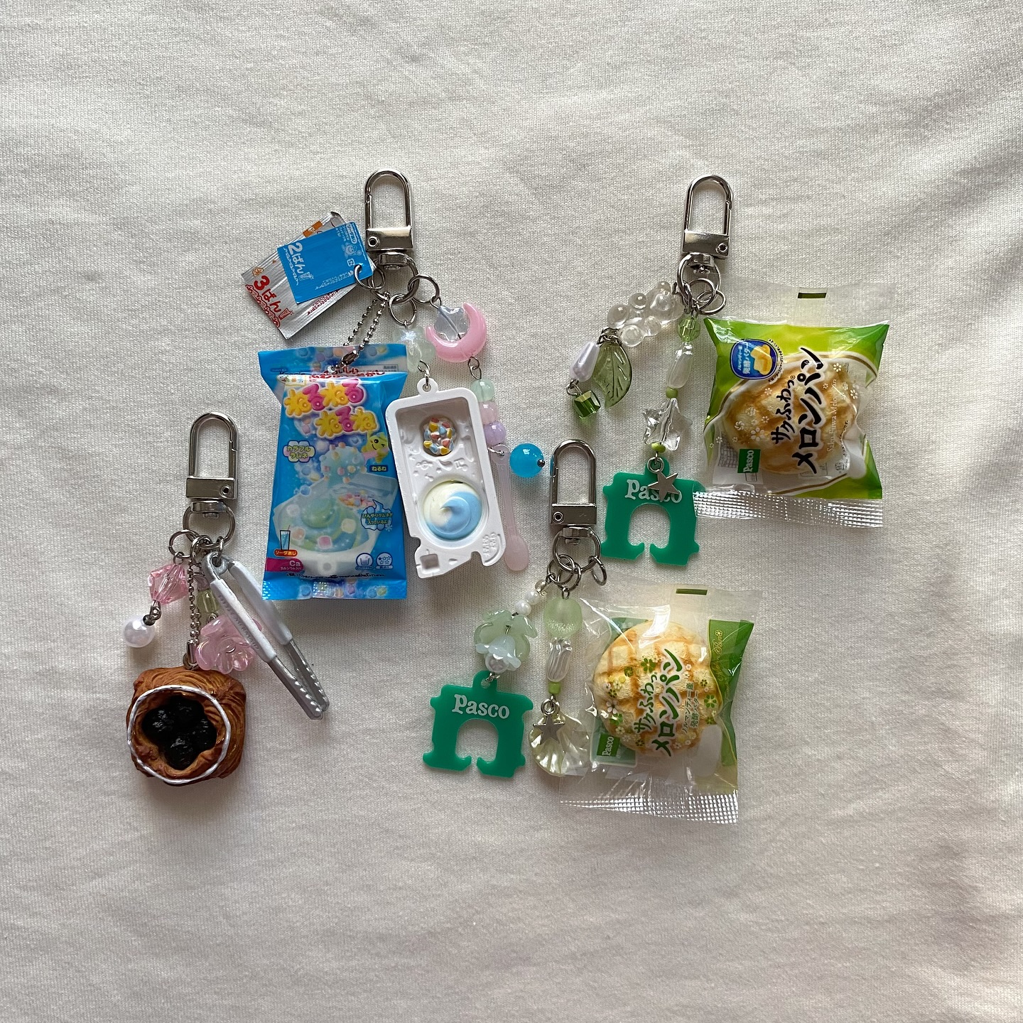 the image showcases four handmade keychains featuring miniature food and candy items. two keychains include miniature bagged melon pan breads, one with a green pasco charm. another keychain features a konapun diy candy kit and a blue candy package. the final keychain shows a boba pastry charm and tiny silver tongs. the keychains are attached with metal clips and embellished with various beads and charms. they are placed on a white, textured surface.