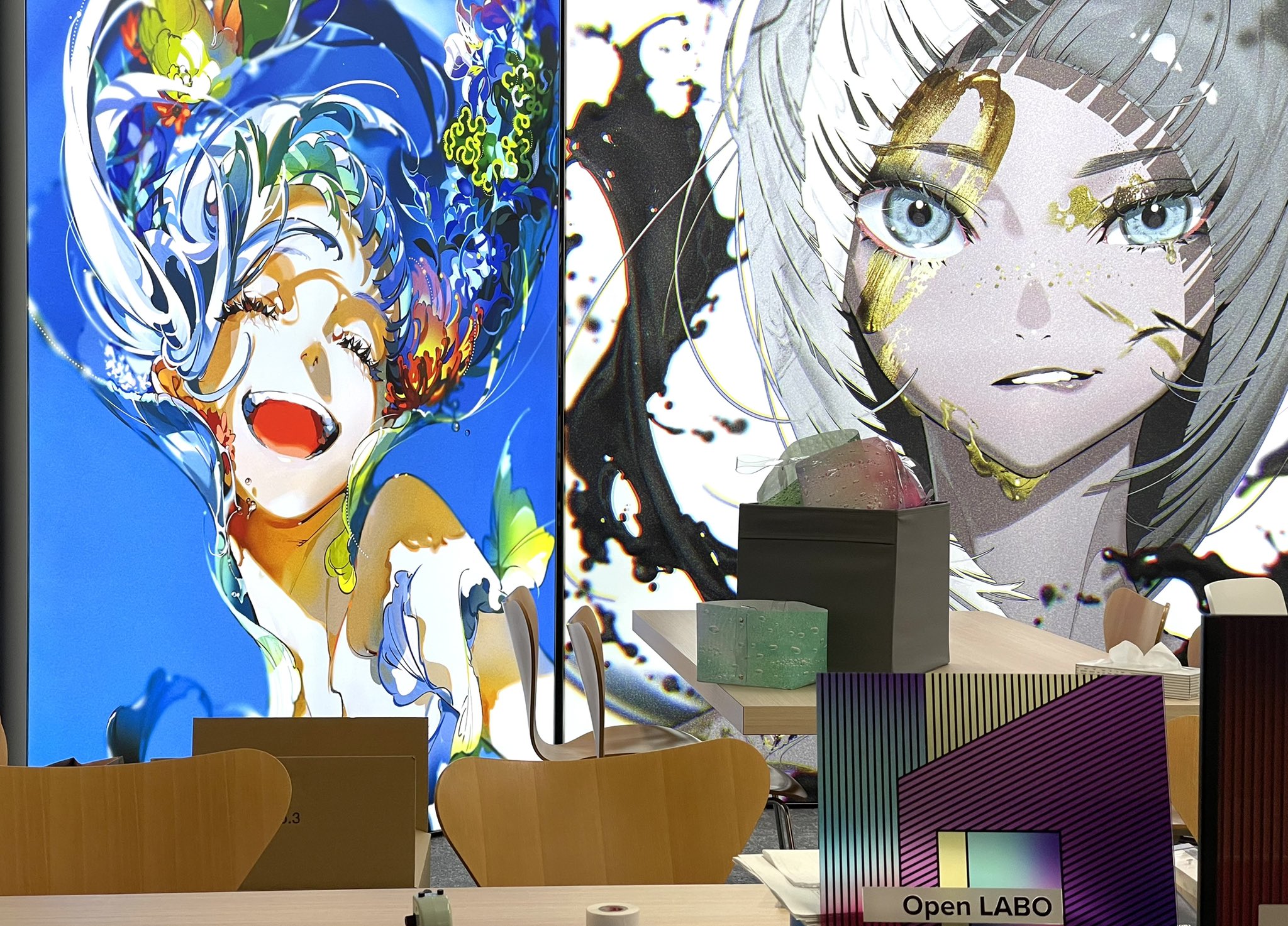 this picture showcases what appears to be an art exhibition or gallery featuring vibrant digital illustrations of animestyle female characters. the artwork utilizes a bold color palette and dynamic compositions. the setting is bright and modern with minimalist furniture, suggesting a contemporary art space.