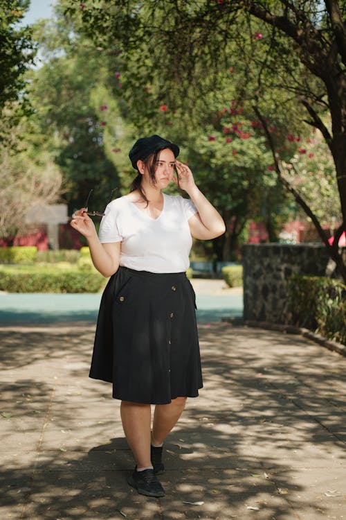 a young woman strolls through a park on a sunny day. she's wearing a white tshirt, black aline midi skirt, black shoes, and a black newsboy cap. she's holding a pair of glasses and has a slightly pensive expression on her face. the scene is peaceful and the colors are muted and natural. the image gives off a casual and relaxed vibe.
