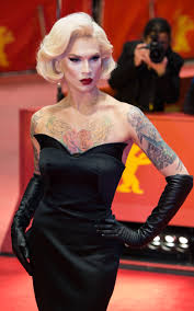a full shot of a drag queen in a black dress with long gloves, posing on a red carpet. she has blonde hair styled in a vintage fashion, and visible tattoos on her arms and chest. she looks directly at the camera. the background is blurry, showing the red carpet and a person taking photos. the image suggests a film festival or similar event.