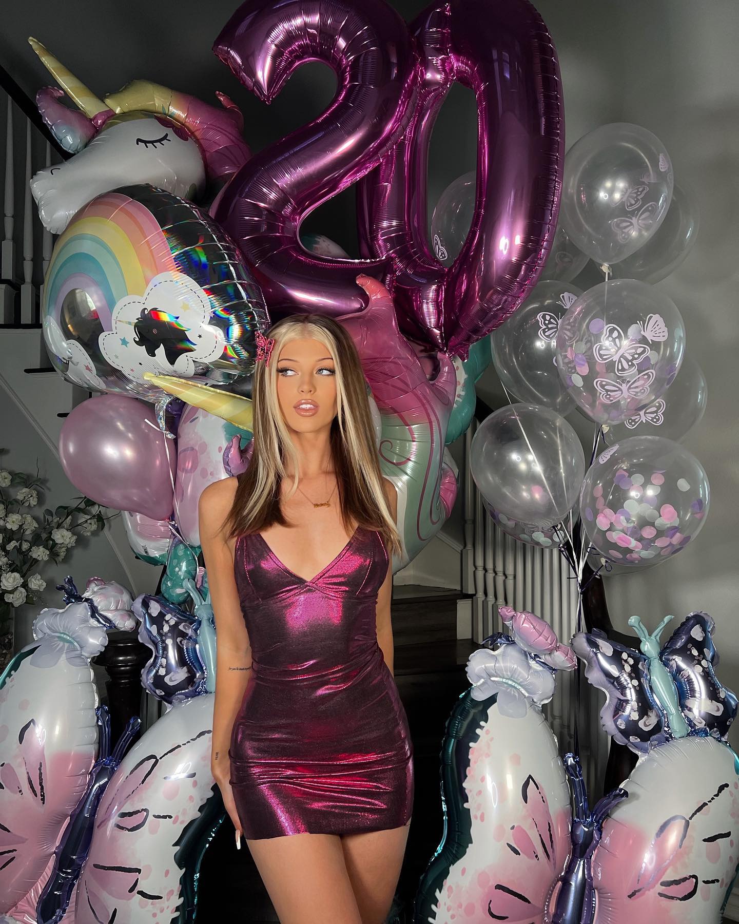 a young woman with blonde hair is standing in front of a backdrop of birthday balloons, including a large "20" balloon, unicorn balloons, and butterfly balloons. she is wearing a shiny, pink mini dress and has a small hair clip in her hair. the background shows a staircase and a decorative floral arrangement. she appears to be celebrating her 20th birthday.