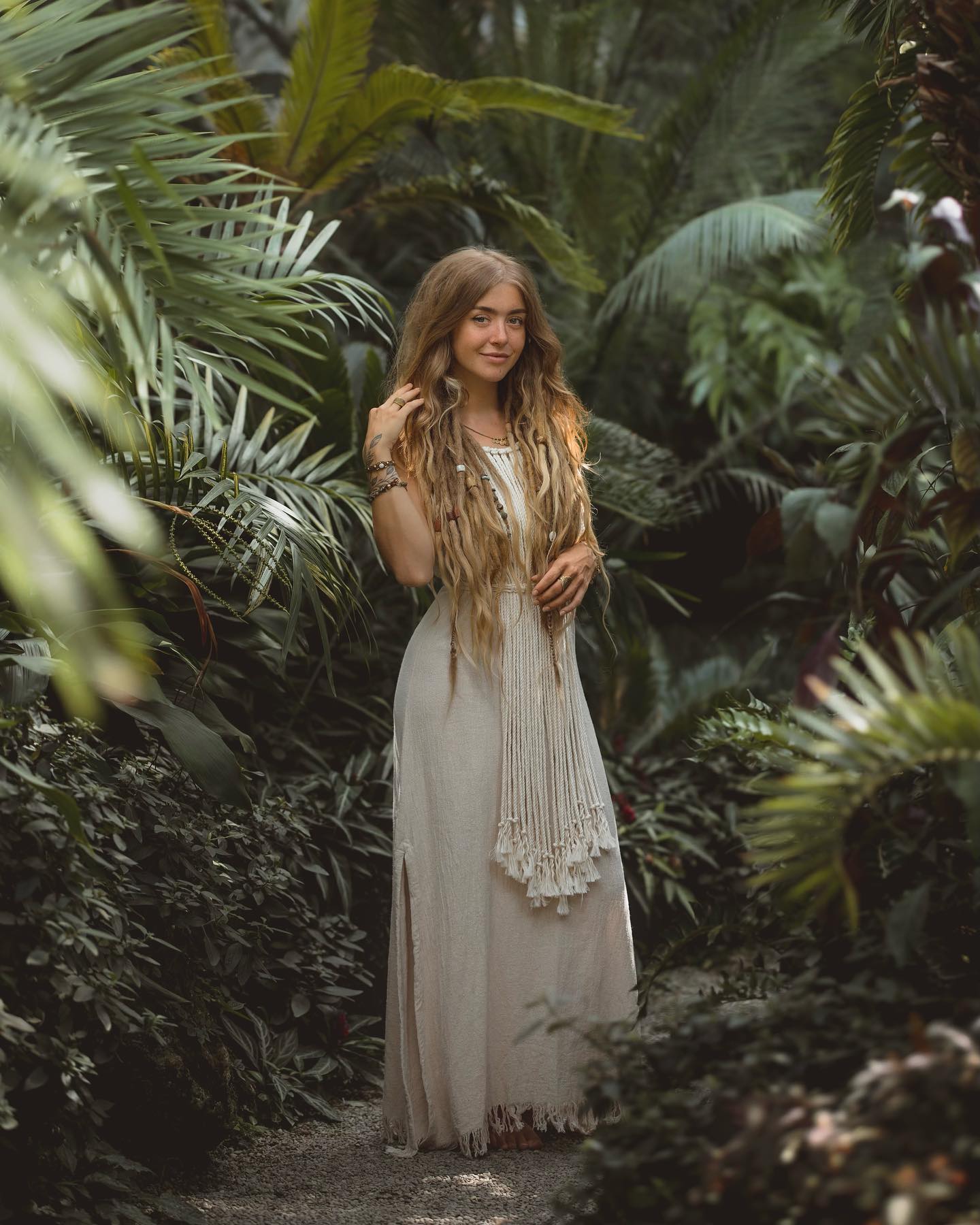 a young blonde woman with long dreadlocks is standing in a lush tropical garden. she is wearing a long, flowing, light beige linen maxi dress with a macrame detail at the waist and fringe at the hem. her attire is bohemian in style. she accessorizes with several bracelets and a macrame necklace. she is smiling gently and appears serene and calm in her environment. the background is filled with dense, vibrant green tropical foliage. the overall mood of the image is peaceful and natural.