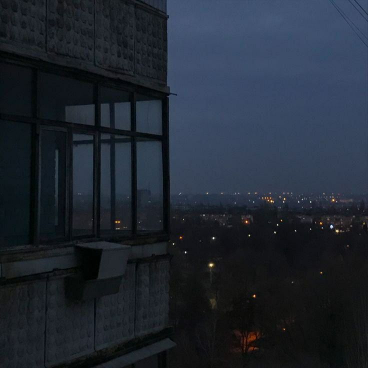 the image shows a highangle, long shot of a city at night. the perspective is from a building's balcony, with a large window taking up a significant portion of the frame. the cityscape is visible in the distance, with numerous streetlights illuminating the scene in a dark and moody ambiance. the building's architecture is plain concrete. the overall mood is quiet, serene, and slightly melancholic. the color palette is dominated by dark blues and greys, emphasizing the night's atmosphere.
