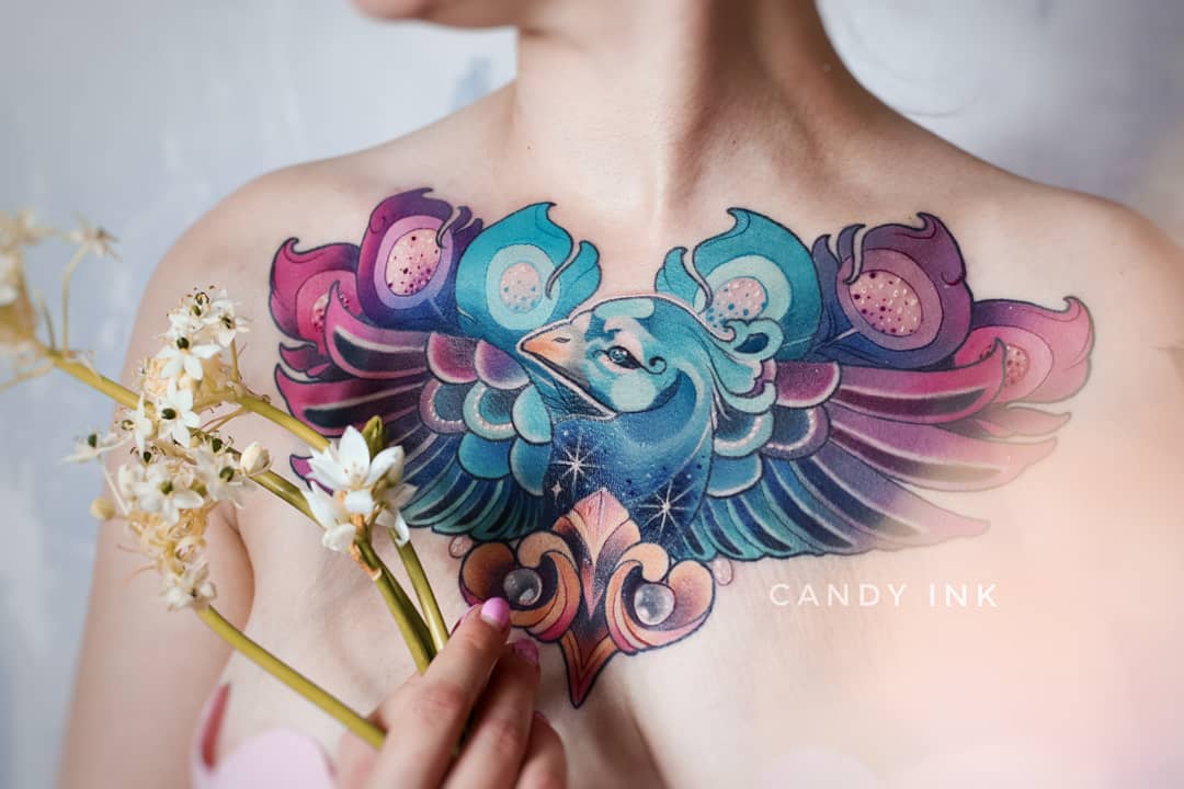 a closeup photograph showcasing a woman's new chest tattoo. the tattoo is of a stylized bird, resembling a cross between a peacock and a phoenix, rendered in vibrant shades of blue, purple, pink, and orange. the bird's wings are spread across the woman's chest, creating a heart shape. delicate floral and geometric patterns embellish the design. the woman holds a small bouquet of white flowers in her hand, positioned near the tattoo. the image is brightly lit, highlighting the intricate details and vibrant colors of the artwork.