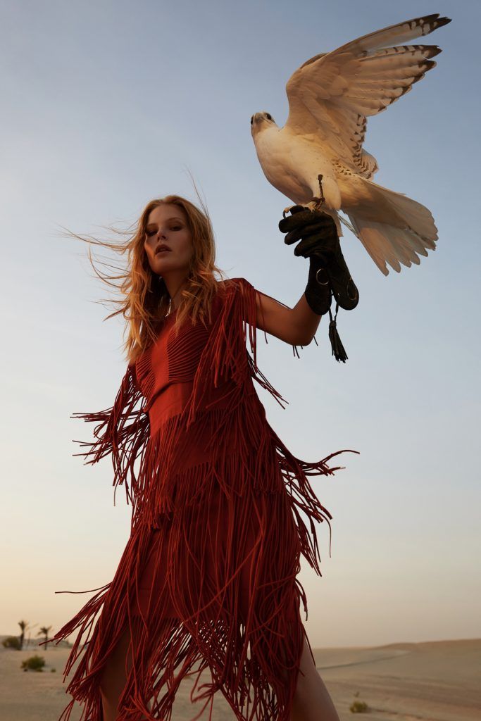 a woman in a red fringed dress holds a falcon in a desert setting. the falcon is white with brown markings, and the woman is wearing leather gloves to hold it. she is looking at the camera with a serious expression on her face. her blonde hair is blowing in the wind. the desert is sandy and the sky is a light blue with some clouds.