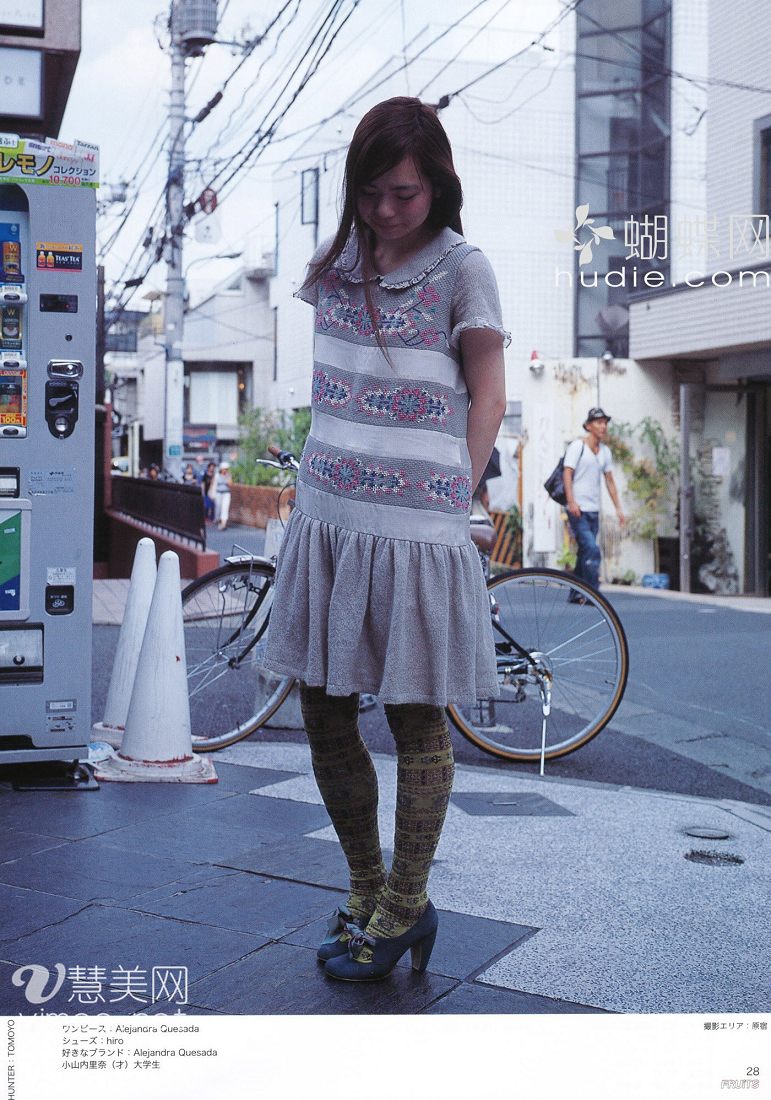 a young japanese woman is captured in a candid street style photograph. she is wearing a unique gray dress with colorful embroidery, paired with patterned tights and blue heels. she stands on a sidewalk with a bicycle leaning against a wall behind her. a vending machine is visible on the left side of the frame, and a man can be seen walking in the background. the overall feel of the image is relaxed and natural.