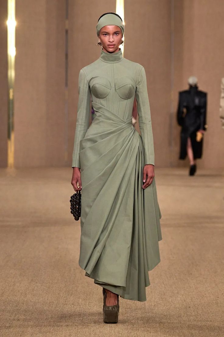 a fashion model walks the runway in a long, green dress with long sleeves and high heels. she is wearing her hair pulled back with a headband and her make up is natural. the runway is beige and the background is blurred. she appears to be holding a small black beaded bag. the model is wearing a corset top with a pleated skirt.