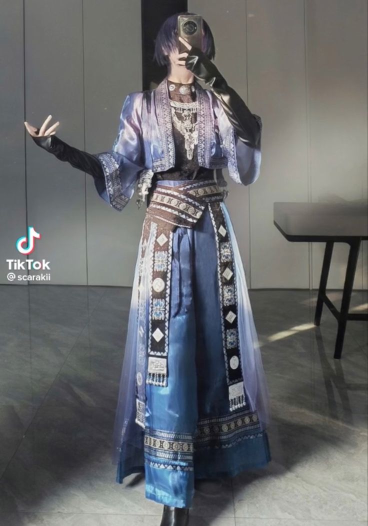 a woman in a blue and white dress with intricate patterns and black accessories poses in front of a mirror. the dress is a flowing, elegant style with a high neckline. the woman has dark hair, styled in a bun. she is wearing a silver necklace and earrings. the backdrop is a white wall with a black table in the foreground.