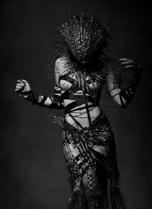 a black and white photo of a woman in a gothic inspired dress. the woman is wearing a unique headpiece that resembles a mask, and the photo appears to be taken in a studio setting. the woman's dress is made of dark, intricate fabric, and she strikes a dramatic pose. the photo is dark and mysterious.