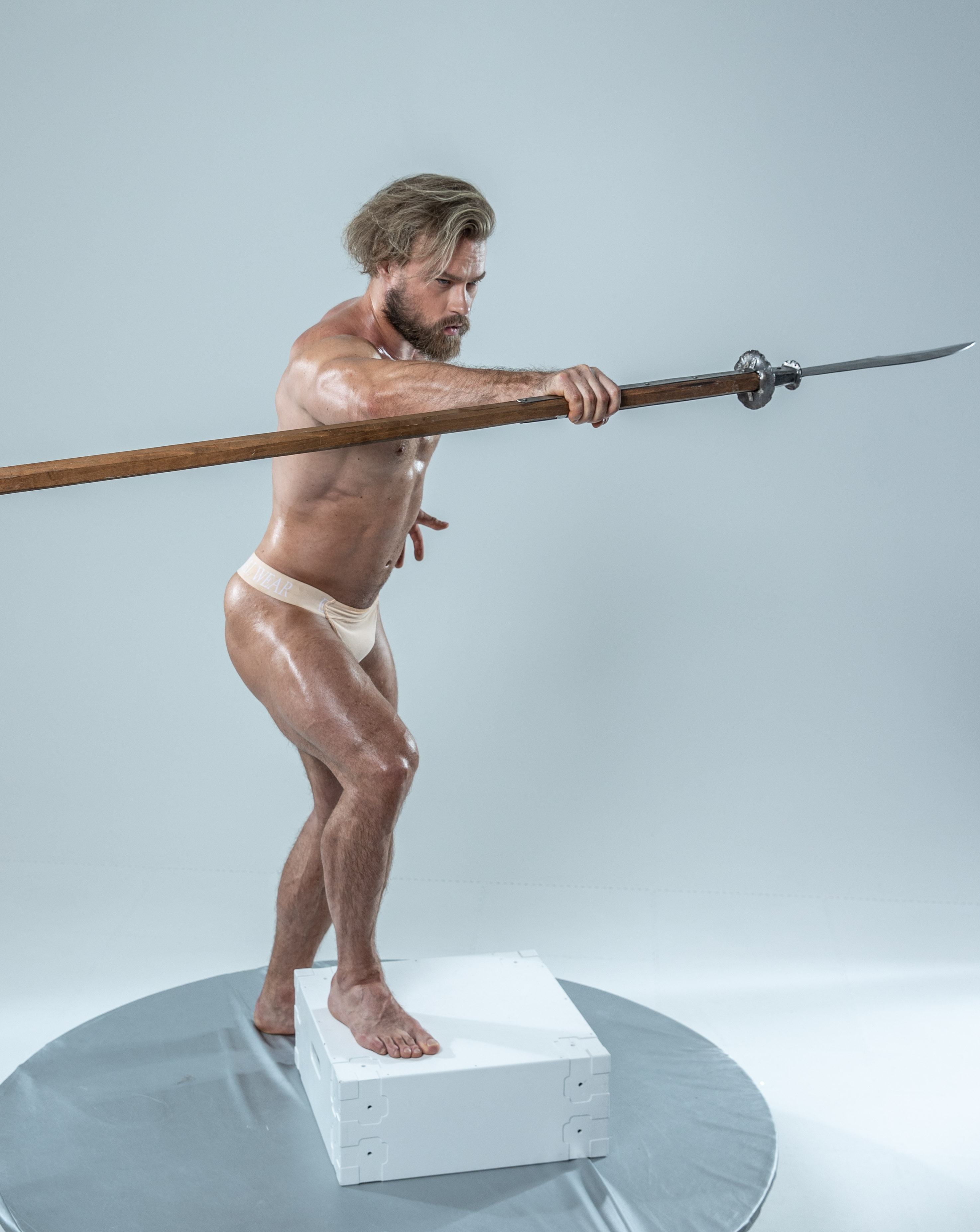 a muscular man with long blond hair and beard is posing in a studio. he's wearing only underwear and holding a spear in both hands, thrusting it forward. he is standing on a white cube, with a grey backdrop behind him. the lighting is bright and even, illuminating his physique.