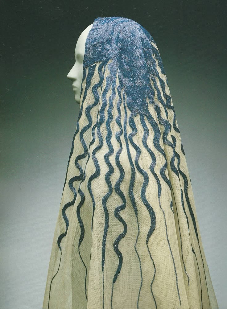 a photograph of a mannequin wearing a creamcolored veil with a blue patterned design. the veil covers the mannequin's head and drapes down, reaching its shoulders. the blue pattern consists of wavy lines that run vertically down the veil. the mannequin is posed in profile against a muted, gradient background.