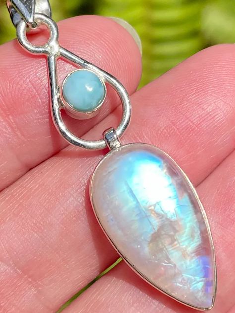 a sterling silver pendant featuring a teardropshaped moonstone and a round larimar cabochon, held in an infinity symbol design. the pendant is displayed against a blurry outdoor background, held by a hand. the natural light enhances the iridescent shimmer of the moonstone.