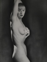 the image is a black and white photograph of a nude woman. she is standing in profile, her body slightly turned away from the camera. her left arm is raised, and her right hand is resting on her hip. her expression is pensive, and her hair is styled in a bob. the lighting is dramatic, with strong shadows creating a sense of depth and texture. the overall style of the photograph is classic and elegant, suggestive of the 1920s or 1930s.